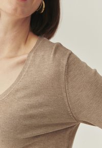 Pull à col en V de couleur marron clair, présentant une coupe ajustée et une texture lisse, avec des coutures visibles le long de l'encolure et des épaules.