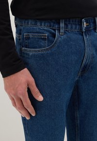 Mano masculina descansando junto al bolsillo de unos vaqueros azules, vistiendo una camisa negra de manga larga.