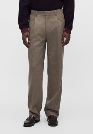 Homme portant un pantalon beige plissé, des mocassins en cuir marron foncé et un haut à manches longues foncé sur un fond clair uni.