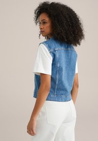 Denimväst i mörkblått med en kort design, som har en krage och bakre yoke, ihop med en vit kortärmad skjorta och vita byxor.