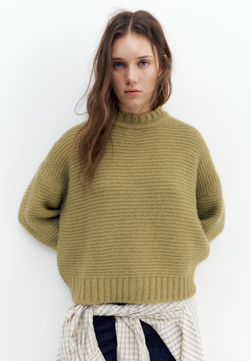 Pull vert en tricot avec un col montant, des épaules tombantes et un ourlet côtelé. Associé à une chemise à carreaux nouée à la taille.