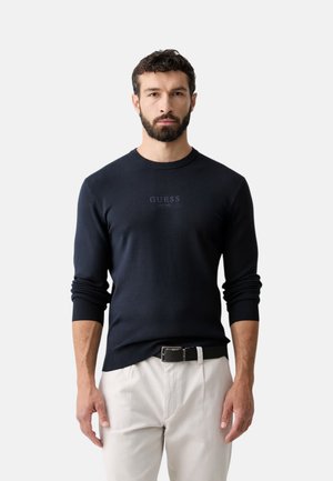 Man met baard die een strak zwart shirt met lange mouwen met het "GUESS" logo draagt en lichtbeige broek tegen een effen achtergrond.