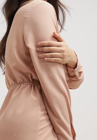 Kvinna i en beige långärmad klänning med samlad midja håller sin överarm med motsatt hand mot en enfärgad bakgrund.