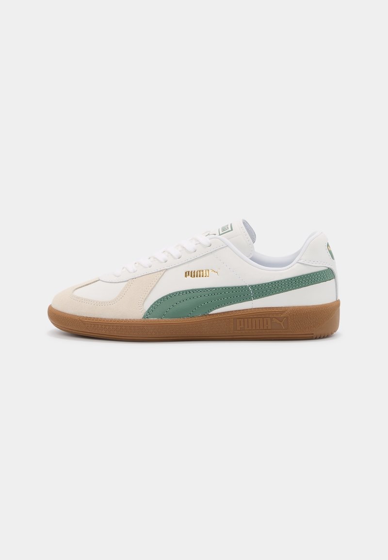 Baskets blanches Puma avec bande latérale verte, empiècement en suède beige, logo Puma doré et semelle en caoutchouc marron sur fond uni.