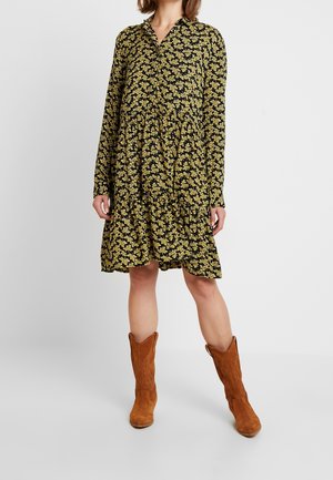 Vestido negro con patrón floral amarillo, mangas largas, cuello y falda ligeramente acampanada. Combinado con botas de vaquero de ante marrón.