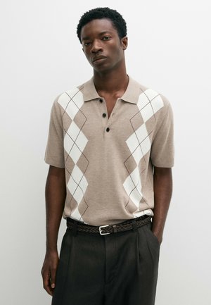 Jonge man die een beige polo met korte mouwen draagt met een wit ruitpatroon en zwarte broek met een gevlochten bruine riem.