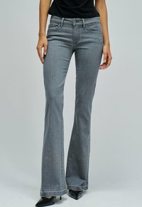 Kvinna som bär grå utsvängda jeans med små rhinestones vid fållen och svarta spetiga klackskor, stående mot en enfärgad bakgrund.