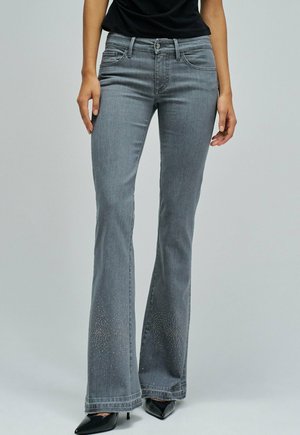 Mujer vistiendo jeans acampanados grises con pequeños detalles de rhinestones cerca de los dobladillos y tacones negros de punta, de pie contra un fondo liso.