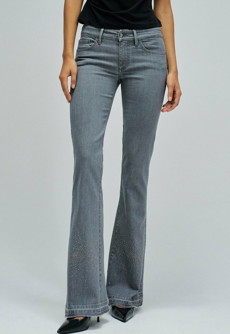 Kvinna som bär grå utsvängda jeans med små rhinestones vid fållen och svarta spetiga klackskor, stående mot en enfärgad bakgrund.