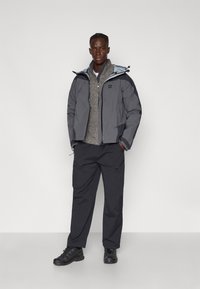 Chaqueta gris impermeable con capucha azul claro, superpuesta sobre un suéter gris. Pantalones oscuros y zapatillas de deporte negras completan el atuendo.