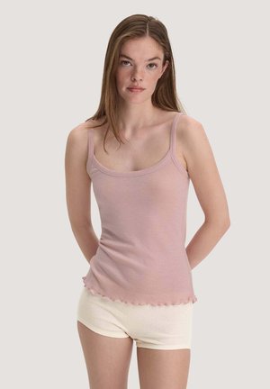 Junge Frau mit langen braunen Haaren, die ein hellrosa Tanktop und cremefarbene Shorts trägt und vor einem einfarbigen Hintergrund steht.