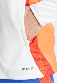 Maglietta sportiva bianca con un pannello laterale a fantasia arancione e rossa, dotata di tasca con zip e logo "AEROREADY" sull'orlo.