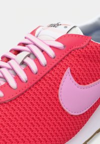 Sapatilhas tricotadas vermelhas com o logo Swoosh rosa, cordões rosa e brancos, e uma língua branca com a marca.