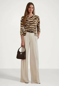 Ženska v majici z leopardjim vzorcem in cardiganom, bež širokih hlačah, drži črno torbico, stoji pred enobarvno ozadjem.