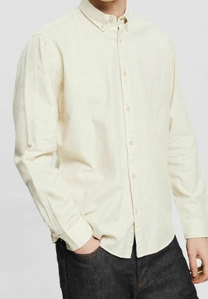 Man wearing a long-sleeve, buttoned, cream-colored shirt with a collar, paired with dark jeans, hand in pocket.