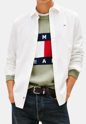 Homme portant une chemise blanche ouverte sur un pull vert avec un motif à blocs rouge, blanc et bleu marine et un jean foncé avec une ceinture marron.