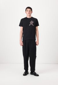 Zwarte katoenen t-shirt met diagonaal wit logo ontwerp, gecombineerd met losse zwarte broek en zwarte schoenen, tegen een effen achtergrond.