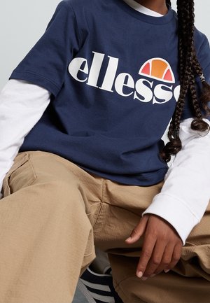 Personne portant un t-shirt ellesse bleu marine par-dessus des manches longues blanches, un pantalon kaki et des baskets noir et blanc, assise avec des tresses visibles.