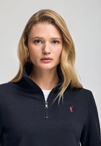 Svart zip-up collegetröja i slät tyg, med en liten röd logotyp på vänster bröst, hög krage och en dragkedja framtill.