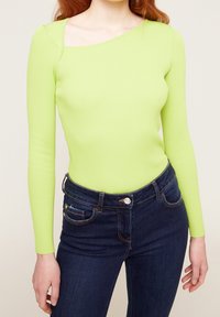 Haut à manches longues en maille côtelée vert lime vif avec un col asymétrique unique, associé à un jean slim bleu foncé avec poches avant.