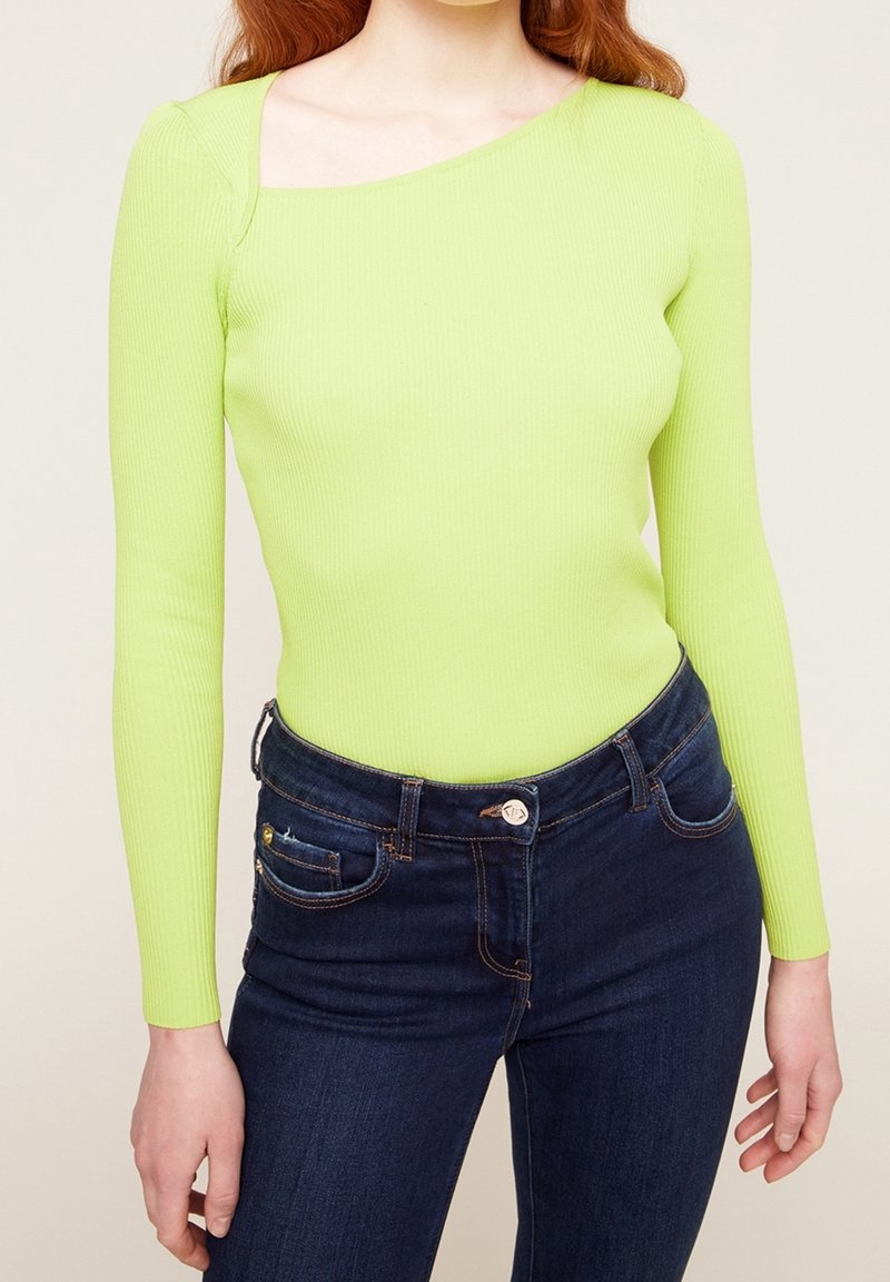 Haut à manches longues en maille côtelée vert lime vif avec un col asymétrique unique, associé à un jean slim bleu foncé avec poches avant.