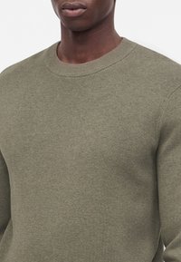Gros plan d'une personne portant un pull en maille vert olive à col rond, montrant le cou, les épaules et le haut de la poitrine sur un fond uni.