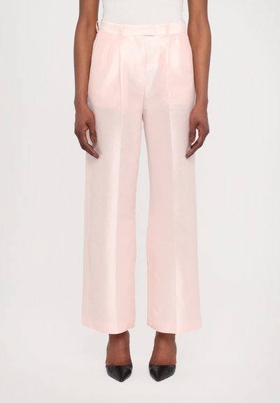 Pantaloni a gamba larga rosa chiaro realizzati in un tessuto lucido, con pieghe frontali e una vita aderente, abbinati a tacchi neri a punta.