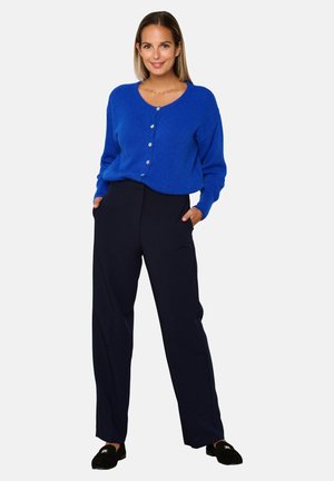 Cardigan blu, oversize, con scollatura rotonda e chiusura a bottoni, abbinato a pantaloni blu scuro. La modella è in piedi con le mani nelle tasche.