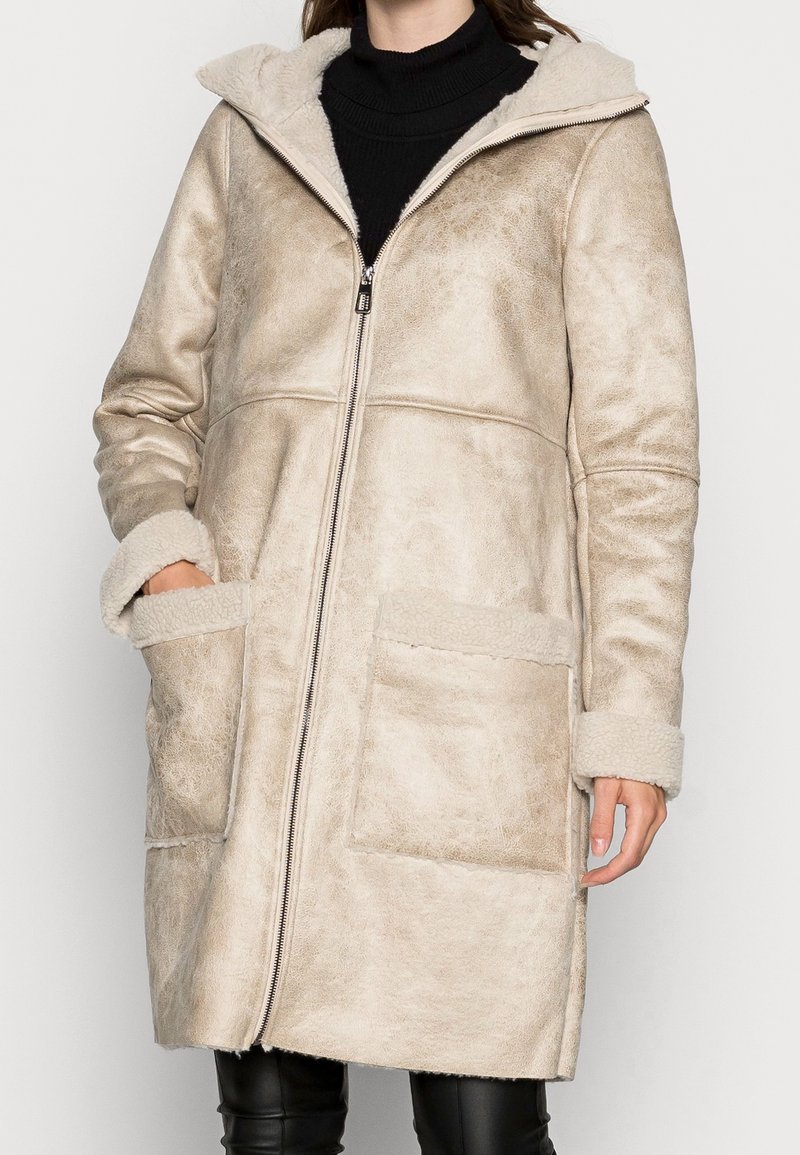 Femme portant un manteau en peau lainée beige à capuche avec de grandes poches avant et une fermeture éclair devant, assorti à un col roulé noir et un pantalon en cuir.
