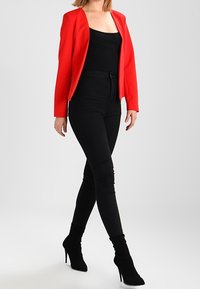 Červený texturovaný blejzr přes černý top, doplněný černými skinny džíny a kotníkovými botami na vysokém podpatku. Oblečení je elegantní a přiléhavé.