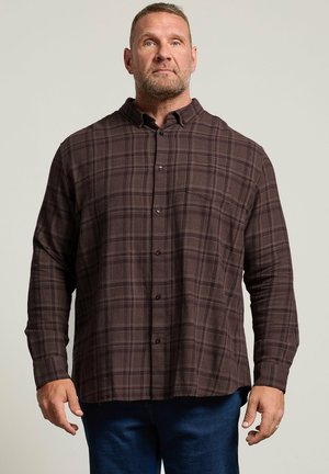Homme aux cheveux courts et à la barbe portant une chemise à carreaux marron foncé boutonnée et un jean bleu, debout devant un fond clair uni.