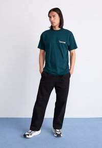 Türkisfarbenes Baumwoll-T-Shirt mit kurzen Ärmeln und einem kleinen Aufdruck auf der Brust. Kombiniert mit schwarzen, lockeren Hosen und gemusterten Turnschuhen.