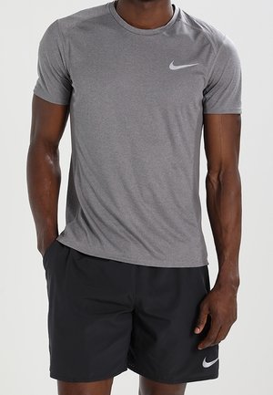 Mann trägt graues Nike-Athletikshirt mit kurzen Ärmeln und schwarze Nike-Shorts, steht mit einer Hand in der Tasche vor einfachem Hintergrund.