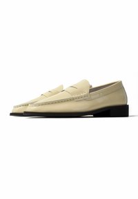 Δερμάτινα loafers σε κρεμ χρώμα με μαύρες σόλες, με κλασικό σχέδιο λουριού penny και χαμηλά τακούνια, εμφανίζονται από πλάγια γωνία σε λευκό φόντο.