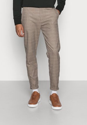 Pantaloni a quadri marroni con una vestibilità slim, dotati di tasche laterali. Abbinati a sneaker marroni e calzini bianchi, su uno sfondo neutro.