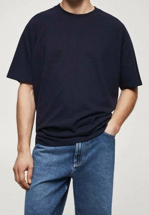 Osoba nosząca gładki czarny T-shirt z krótkim rękawem i niebieskie jeansy, stojąca z jedną ręką w kieszeni, a drugą opuszczoną swobodnie wzdłuż ciała.