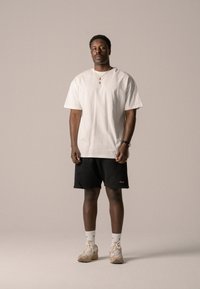 Weißes Baumwoll-T-Shirt, kurze Ärmel, lässige Passform; kombiniert mit schwarzen Shorts, goldene Logodetails; hellbeige Sneakers; schlichte weiße Socken.