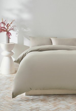 Neutrales beiges Bett mit passenden Kissen und Bettdecke in einem minimalistischen Schlafzimmer neben einem skulpturalen weißen Beistelltisch mit einer Vase mit rotem Laub.
