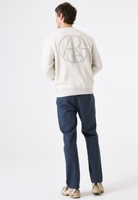 Lichtgrijze sweatshirt met een groot, gestructureerd cirkelvormig logo op de achterkant, gecombineerd met marineblauwe broeken en tan sneakers.
