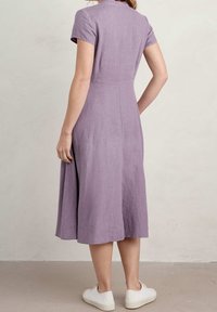 Kvinna som vänder bort, klädd i en knälång, kortärmad lavendelfärgad klänning och vita sneakers, stående på ett enfärgat golv mot en ljus vägg.