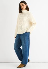 Chunky cream cable-knit turtleneck sweater with dropped shoulders, paired with high-waisted blue wide-leg jeans and tan suede ankle boots.