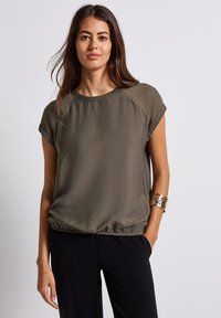 Blusa a maniche corte di colore verde oliva realizzata in tessuto leggero, con scollatura rotonda e orlo elasticizzato. Modella che indossa un braccialetto decorativo.