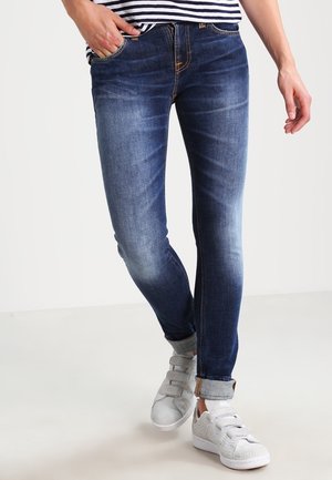 Persoon draagt slim-fit blauwe spijkerbroek met omslagen en witte sneakers, met één hand in de zak en een gedeeltelijk zichtbare gestreepte top.