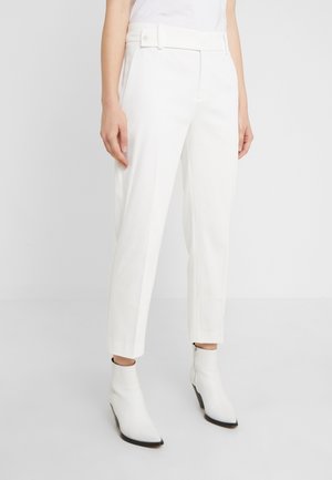 Vrouw die een witte op maat gemaakte broek met hoge taille draagt, met geknoopte riemlussen en witte enkellaarzen met een puntige neus, staand tegen een effen achtergrond.