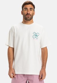Hvid bomulds t-shirt med et teal- og pinkgrafikdesign på venstre bryst. Korte ærmer og en afslappet pasform.
