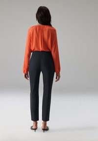 Fel oranje blouse met een geplooide halslijn en lange mouwen, gecombineerd met strakke zwarte broek. Gladde stof en slanke pasvorm.