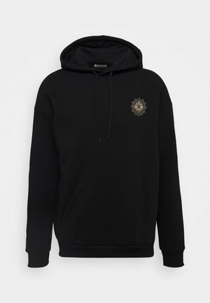 Svart hettegenser laget av mykt stoff, med en frontlomme og et brodert gullløveemblem på venstre bryst.