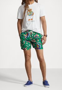 Wit T-shirt met een berenafbeelding, gecombineerd met groene zwembroek met bloemen- en nautische patronen, gedragen met blauwe sneakers.