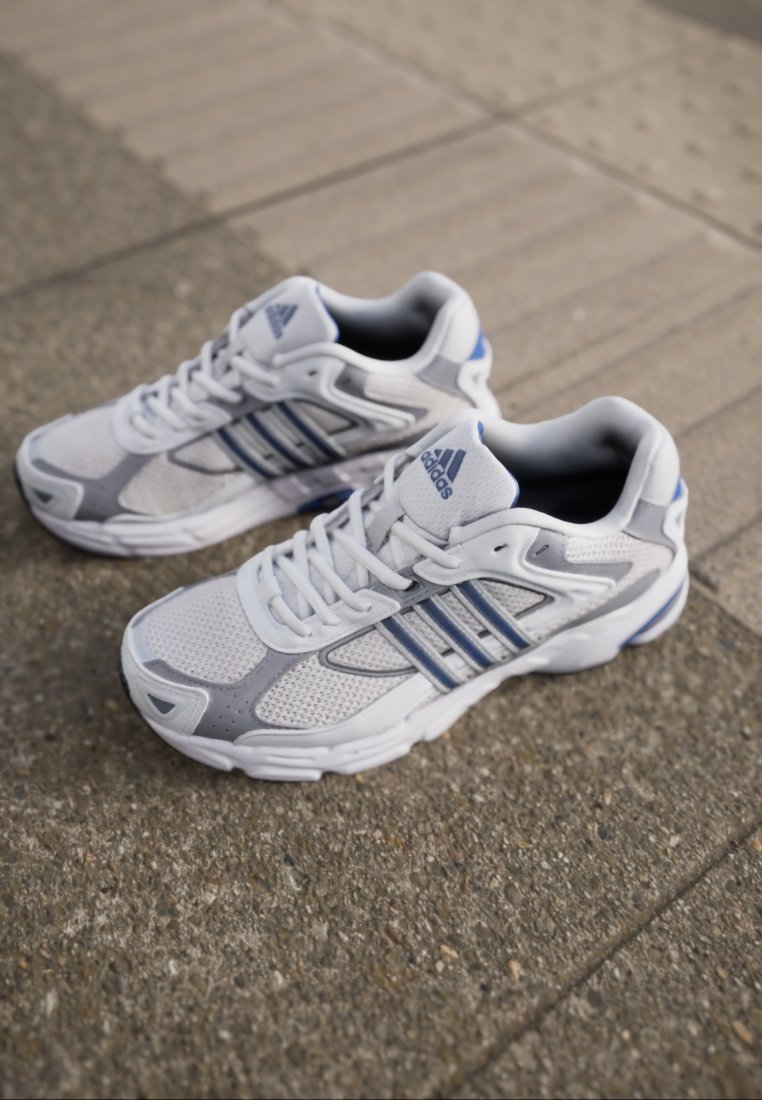 Zapatillas deportivas blancas con detalles en gris y azul, material de malla, puntera redondeada, suela gruesa y logo de tres rayas en el costado.