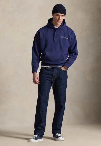 Mladý muž v tmavomodrej čiapke zvaný beanie, tmavomodrej mikine s logom, tmavých džínsoch, teniskách a s hodinkami na zápästí, pózujúci pred jednofarebným béžovým pozadím.